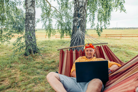 man relaxing in hammock working on laptop in nature in red cap and yellow shirt with green grass background, remote work travel blog digital marketing outdoor lifestyle, copy spaceの写真素材