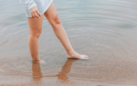 womans barefoot legs on wet sandy beach with feet in shallow water at lake shore wearing short white dress concept of wellness travel summer retreat fashion lifestyleの写真素材