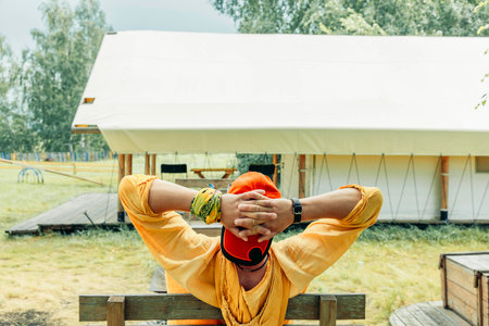 man in orange cap and yellow hoodie leaning back on wooden bench with hands behind head in front of glamping tent in forest meadow on summer day, travel eco tourism slow lifestyle retreatの写真素材