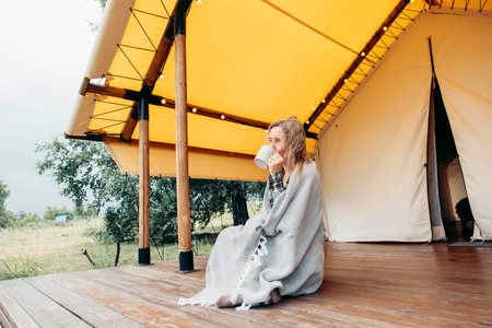 woman drinking from white mug while sitting on wooden terrace of a glamping tent wrapped in blanket...
