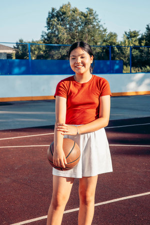 smiling Asian teenage girl in red shirt, white skirt holding basketball on outdoor court on sunny summer day, youth sport girl empowerment healthy lifestyle and leisure physical activityの写真素材