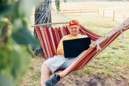 man relaxing in hammock working on laptop in nature in red cap and yellow shirt with green grass background, remote work travel blog digital marketing outdoor lifestyleの写真素材