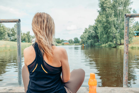 close up woman sitting cross-legged on wooden dock over calm river, wearing sportswear, back view, summer greenery and countryside houses, meditation, morning ritual, nature therapyの写真素材