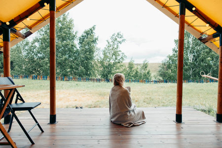 Unrecognizable woman wrapped in patterned blanket sitting on glamping terrace and gazing at lush...