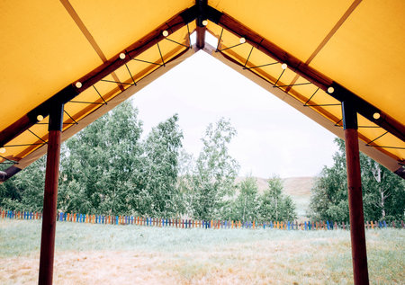 A spacious glamping tent with a yellow canopy. The view shows green trees and a wooden fence...