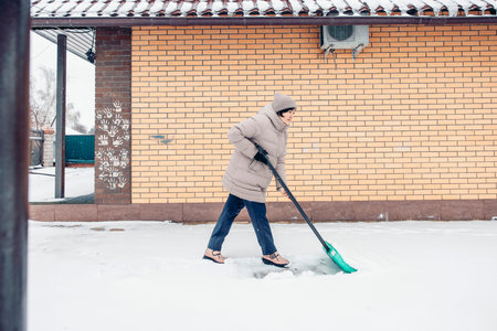 A young Asian woman in a beige winter coat shovels snow outside a brick house, while clearing the snow.の写真素材