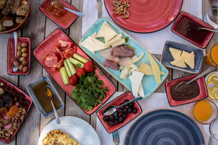breakfast prepared on wooden table with cheeses, jams, tomatoes, cucumber, smoked meat, butter, honey, olives, breadsの写真素材