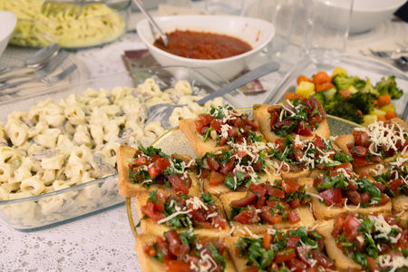 A beautifully set table with a homemade Italian-inspired feast, featuring crispy bruschetta topped with fresh tomatoes, herbs, and cheese, a creamy tortellini pasta dish with mushrooms, a bowl of rich marinara sauce, and a side of roasted vegetables including broccoli, carrots, and potatoes. The elegant presentation on a lace tablecloth adds warmth and authenticity to the dining experience. Perfect for showcasing home-cooked meals, traditional Italian cuisine, and gourmet dining.の写真素材