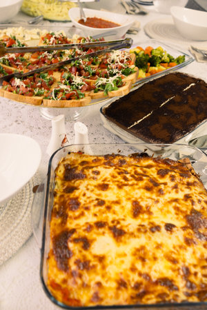 A beautifully arranged homemade Italian dinner featuring a freshly baked lasagna with golden-brown melted cheese, crispy bruschetta topped with diced tomatoes, herbs, and cheese, a rich tiramisu dessert, and a side of roasted vegetables. The dishes are set on a lace tablecloth, creating a cozy and inviting dining atmosphere. Perfect for food blogs, restaurant promotions, or home-cooked meal inspirations.の写真素材