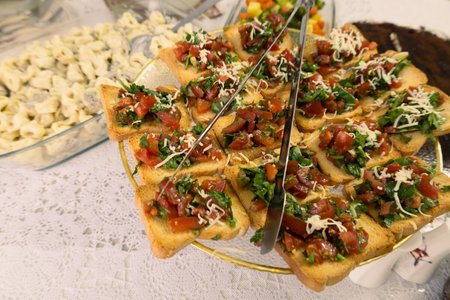 A close-up of a beautifully arranged platter of homemade bruschetta, topped with fresh diced tomatoes, herbs, and shredded cheese on crispy toasted bread. The dish is served on an elegant gold stand over a lace tablecloth, creating a warm and inviting dining atmosphere. In the background, creamy tortellini paste and a variety of colorful vegetables add to the rich Italian-inspired meal. Perfect for food blogs, restaurant promotions, and culinary inspirations.の写真素材