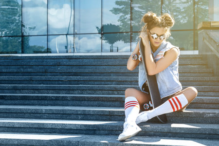 Stylish girl with a skateboard sitting on the stairs in city center