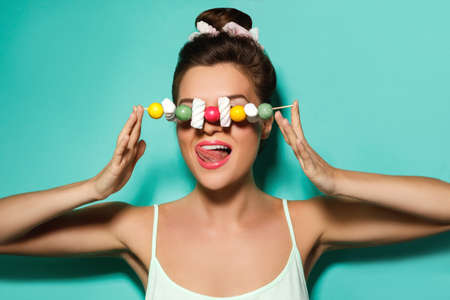 Happy woman with colorful makeup and sweet candies on skewer against cyan backgroundの写真素材