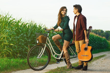 Young couple is walking by country road. Girl riding bicycle and guy is holding guitar.