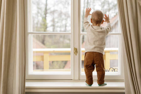 Baby boy is standing on the windowsill and looking at the windowの写真素材