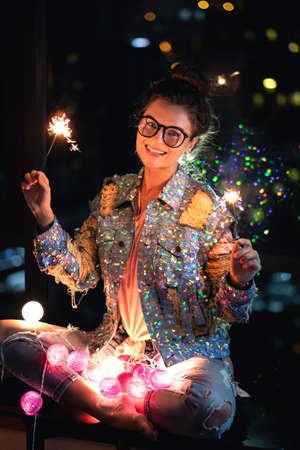 Happy woman wearing glowing jacket with sequins is holding sparklers in her hands.の写真素材