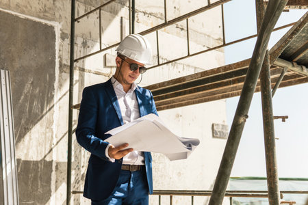 Architect wearing formal suit and hard hatholding a blueprints on a construction siteの写真素材