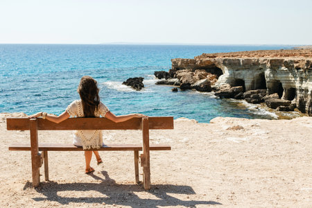 Woman sits on a bench near sea cliffs at Cape Greco viewpoint, enjoying the scenery and natural beauty.の写真素材