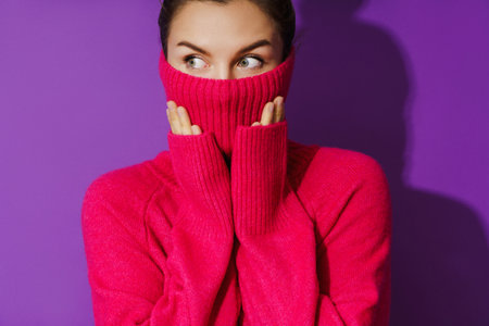 Young woman is hiding her face inside a warm and cozy polo neck sweater against purple backgroundの写真素材