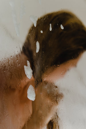 Woman is washing her hair with a shampoo while taking shower. Focus on the wet glass.の写真素材