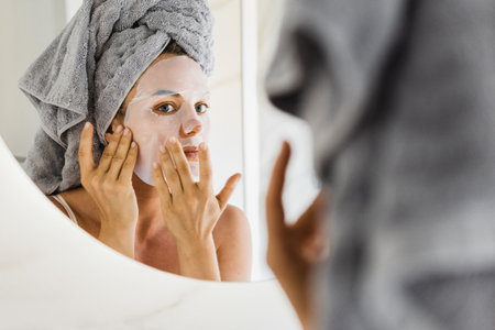 Woman in bathroom with applied sheet mask on her face looking in the mirrorの写真素材