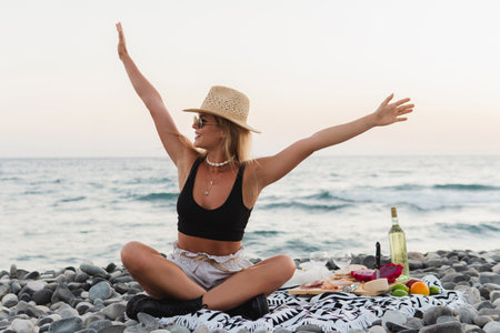 Stylish and carefree woman wearing a straw hat is having a serene picnic on a pebble beach during a sunsetの写真素材
