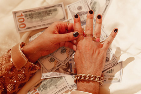 Hands of a wealthy man, adorned with golden rings and a striking black manicure against a backdrop of US dollar billsの写真素材