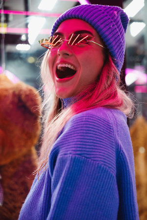 Cheerful and stylish woman, dressed in warm clothes and wearing sunglasses shaped like fire flames, is having fun in a snowy winter amusement parkの写真素材