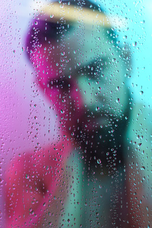 Portrait of young man captured through wet glass in colorful lightの写真素材
