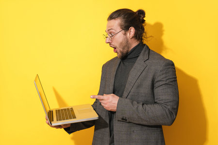 Surprised bearded man wearing eyeglasses pointing at laptop screen on yellow backgroundの写真素材