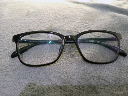 A pair of black-framed eyeglasses rests neatly on a plush grey fabric surface, reflecting light with a slight green hueの写真素材