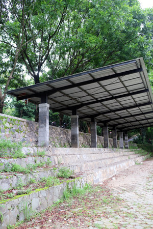A tiered stone seating area covered with a modern metal roof structure supported by concrete pillars, set in a green park environmentの写真素材