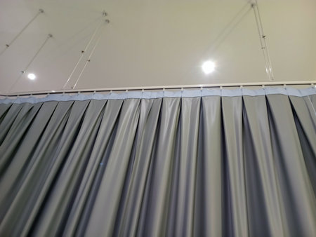 A close up view of a gray curtain with pleats, hanging from a ceiling track. The curtain fabric appears smooth and shiny, with soft lighting illuminating the scene from above.の写真素材