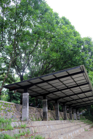 A modern pavilion with a metal roof supported by stone columns, surrounded by lush green trees. The structure is situated in a natural setting, providing shade and a place for relaxationの写真素材