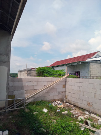 A view of a neglected outdoor area with a concrete wall, overgrown grass, and scattered debris. In the background, there are two buildings with a red roof and clothes hanging outsideの写真素材