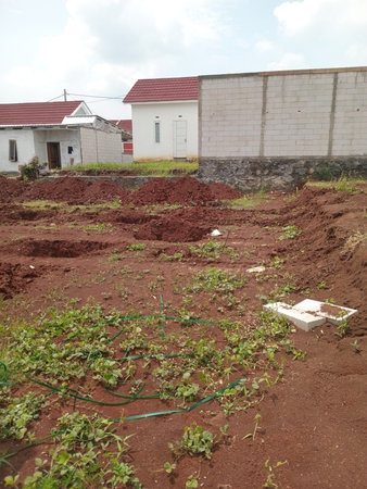 A construction site with red soil, marked foundations, and scattered vegetation. Nearby structures include a white house and a stone wall. The sky is partly cloudy.の写真素材