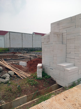 A construction site featuring stacked white building blocks, a foundation pit, wooden planks, and a backdrop of unfinished walls. The area is surrounded by greenery and has a cloudy sky.の写真素材
