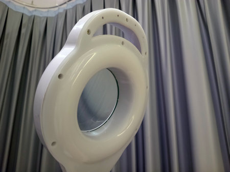 A close up view of a white magnifying lamp with a circular lens, designed for detailed work or examination. The background features soft, draped curtains in shades of gray.の写真素材