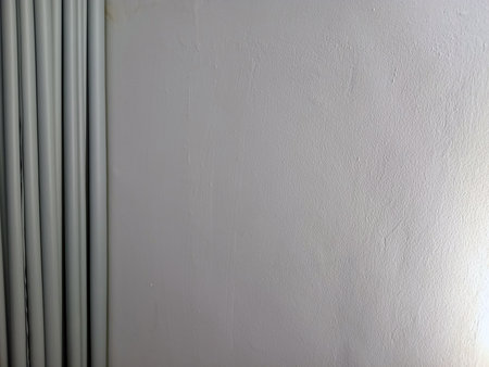 A close up view of a textured white wall with subtle imperfections and light reflections. The image also includes vertical lines of light colored pipes on the left side.の写真素材