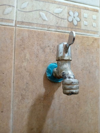 A close up of a rusty water tap mounted on a tiled wall. The tap has a blue tape around its base, indicating a temporary fix. The tiles feature a floral design.の写真素材