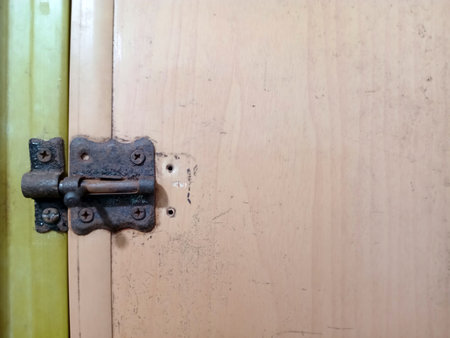 A close up view of a rusty door hinge on a wooden door. The hinge shows signs of wear and age, with a light colored wooden surface and a partially visible latch.の写真素材