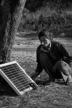 Black and white street photograph of man charging his mobile device from the solar panel while sitting down and staring towards the camera in the month of January 2019 in Dehradun, Uttarakhand, Indiaのeditorial素材