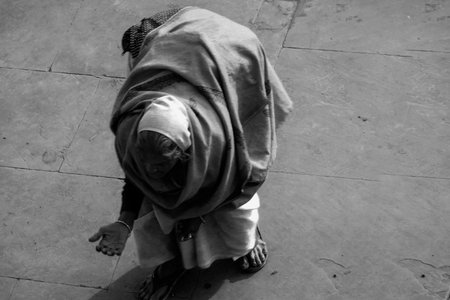Black and white street photograph of an old lady with crooked spine and no visible face begging on...