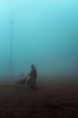 Old woman sweeping the street with a broom in a foggy morning