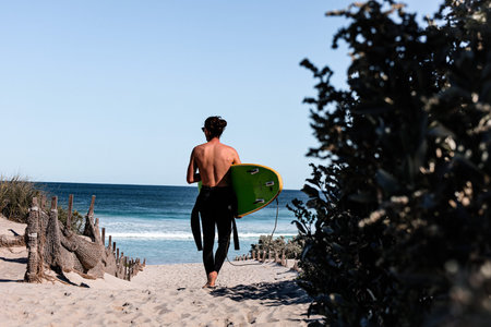 Surfer with wetsuit getting ready for a surf session. Boy walking on the beach with a green...