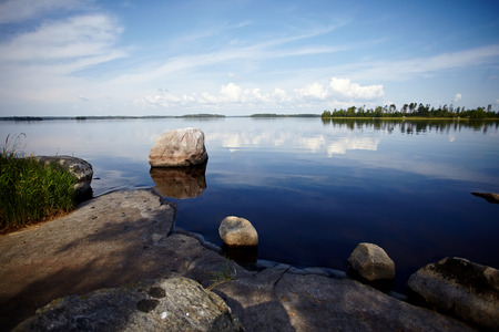 Water landscape with stones. Stones in water. The lake with stones. Beautiful landscape. Water smooth surface and the blue sky with clouds. Stone coast of the lake.の写真素材