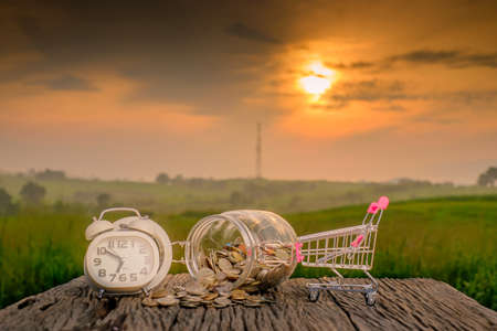Mini supermarket shopping cart , white alarm clock and coins in the jar on old wood with blurred green nature background and beautiful sunrise.の写真素材