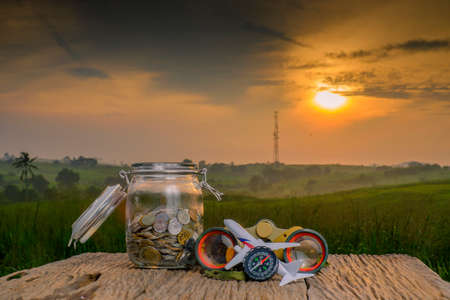 Saving planning for Travel budget of holiday concept,Financial, binocular, coins money in the jar, compass and airplane wooden table with sunrise backgroundの写真素材