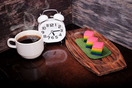 Traditional Malaysian food. Kuih Lapis , alarm clock and  a cup of coffee on wooden background. Coffee time conceptの写真素材