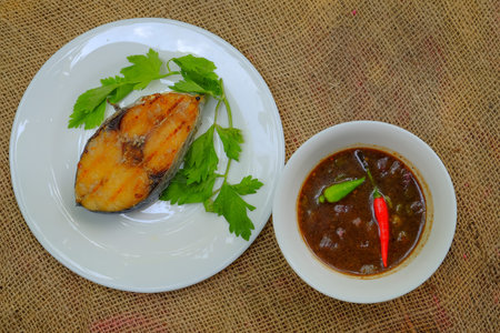 A grilled mackerel fish on white plate served with tamarind sauce is a one of the Malaysian people...