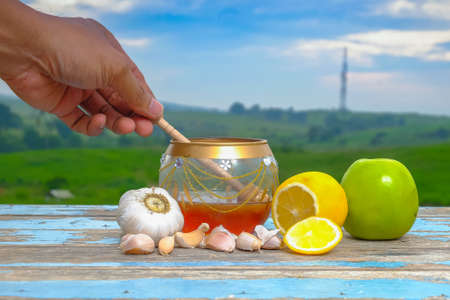 Honey dripping from a wooden honey dipper in a jar. Fresh honey in glass jar , garlic, lemon and green apple on old wooden with blurred background , healthy nutrition, strengthening immunity and treatment of fluの写真素材
